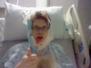 Post Op Me, bloodied in bandages in a hospital bed, giving a thumbs up, immediately after jaw surgery.